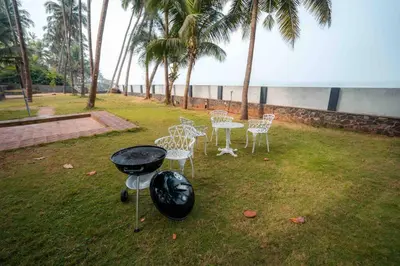 a grill and chairs in a yard with palm trees at Baywatch by Grha - A Picturesque Beachfront Villa in Kozhikode