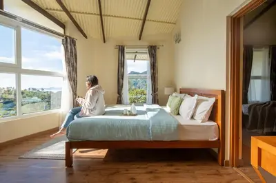 a woman sitting on a bed looking out the window at SaffronStays Valleyfall in Kotagiri