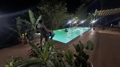a man sitting in a chair by a swimming pool at night at Golden Cypress Resort with Pool -Wayanad in Padinjarathara